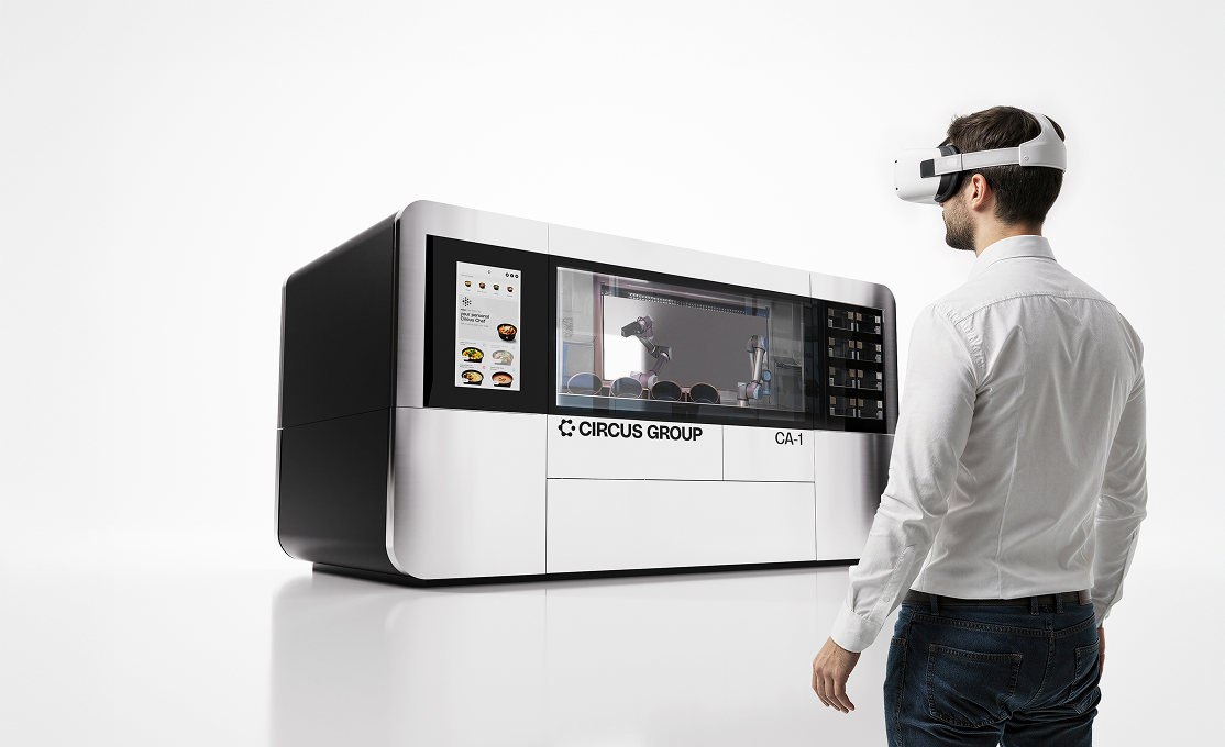 Man wearing a white shirt and VR headset stands in front of a large modern machine labeled Circus Group CA-1 with robotic arms inside.