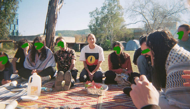 A group of people sitting in a circle outside on a pattern mat. Leah House is seated in the middle. Some people are wearing Keffiyehs and there is a tray of sliced watermelon in the centre.