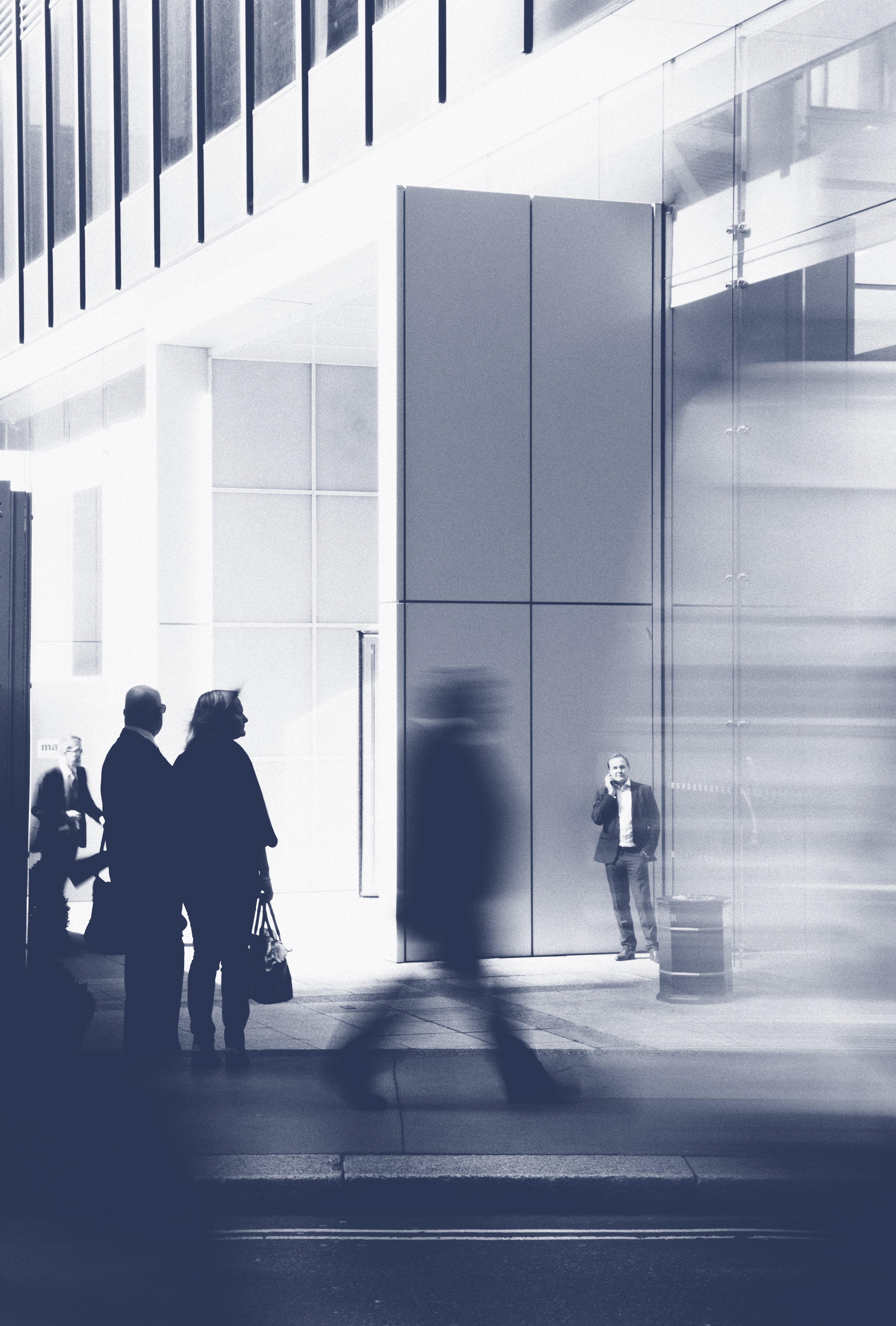 Silhouettes of people walking and standing near a modern building with large glass windows.