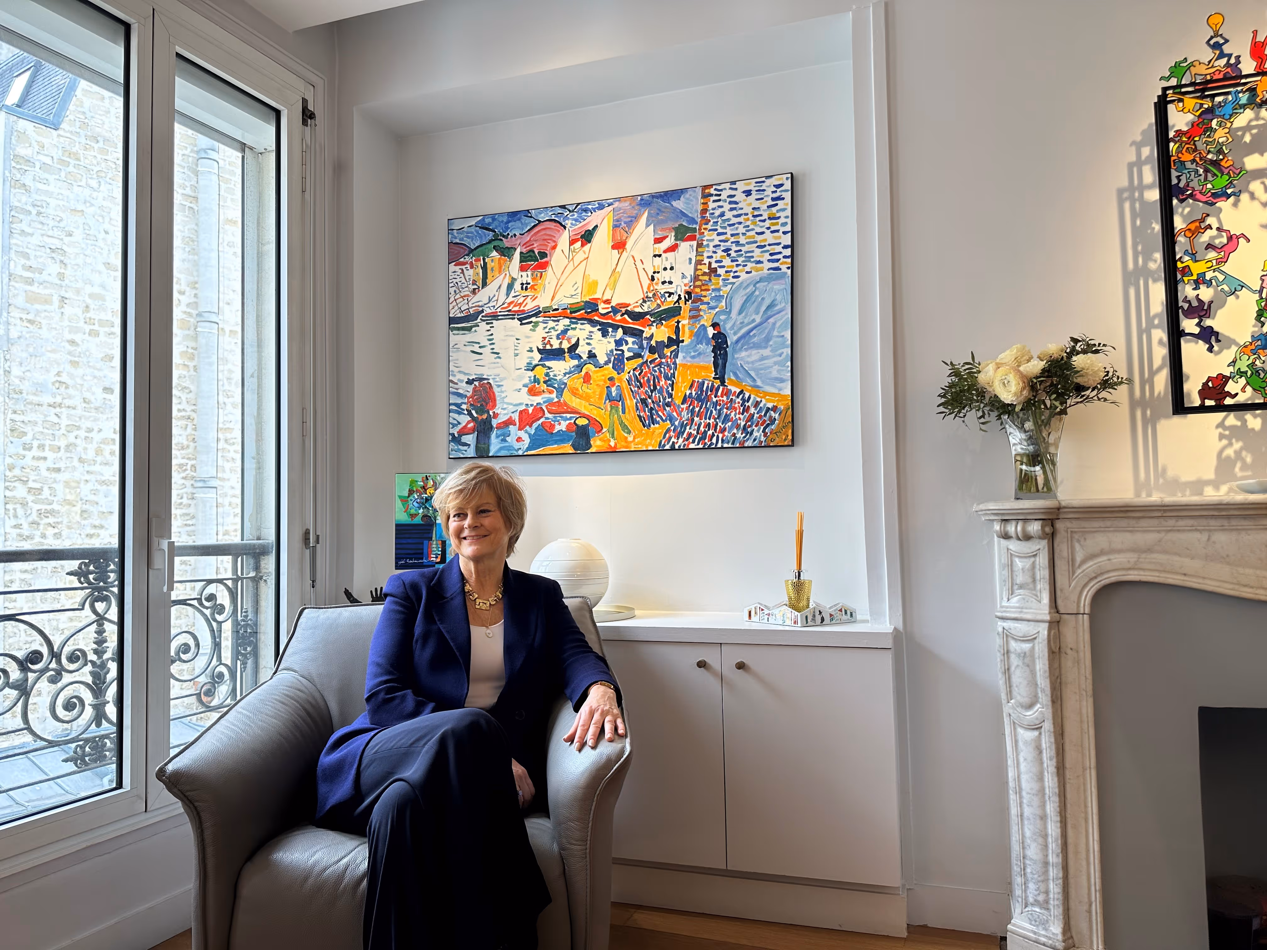 Woman in a navy blazer sitting on a gray armchair in a bright room with colorful paintings, a flower vase on a marble fireplace, and a large window with wrought iron railing.