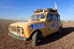 Yellow vintage rally car with roof rack parked on dry, cracked earth under a blue sky.