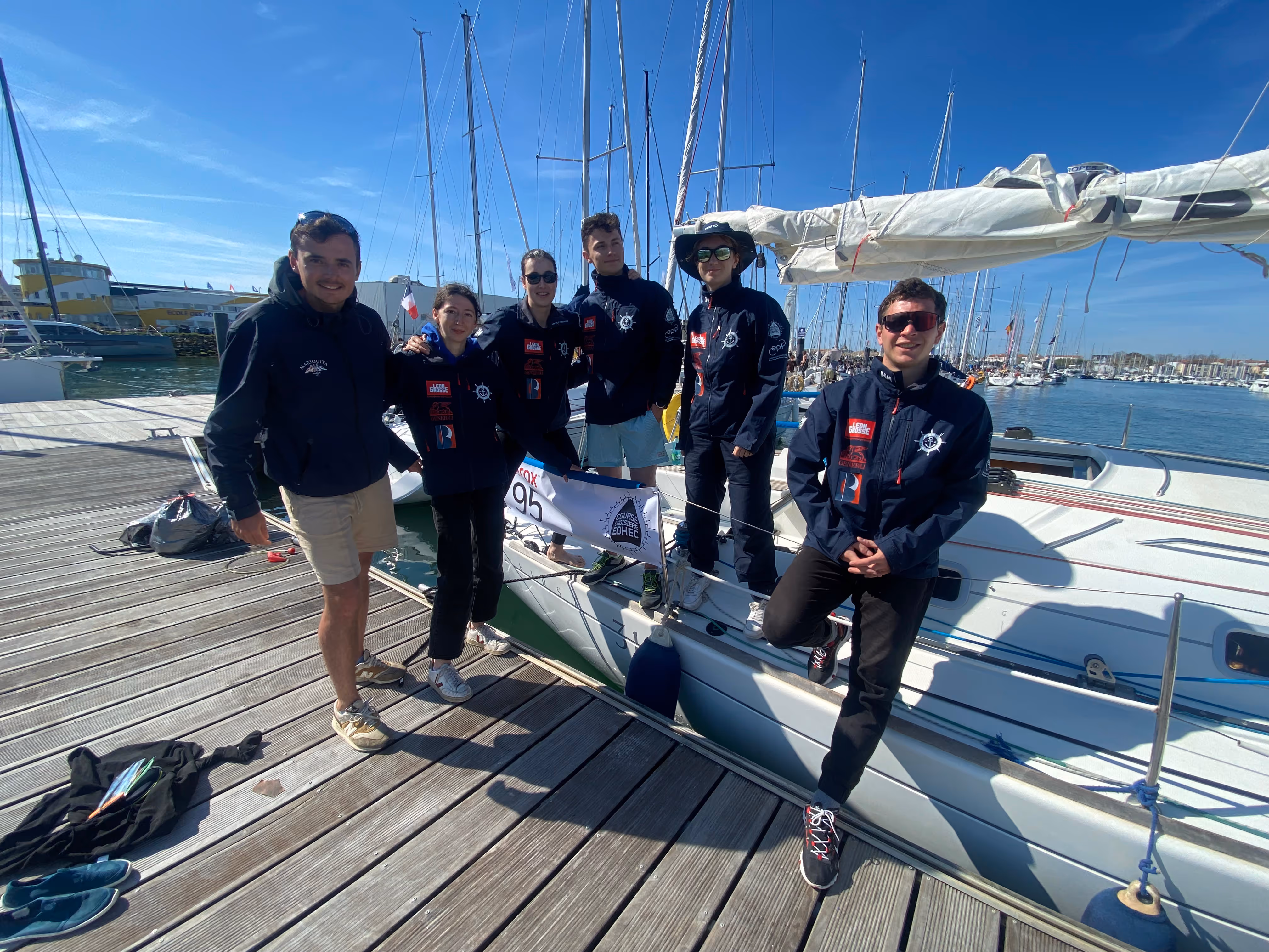 Group of six young people in sailing jackets posing on a dock and a sailboat under clear blue sky at a marina.