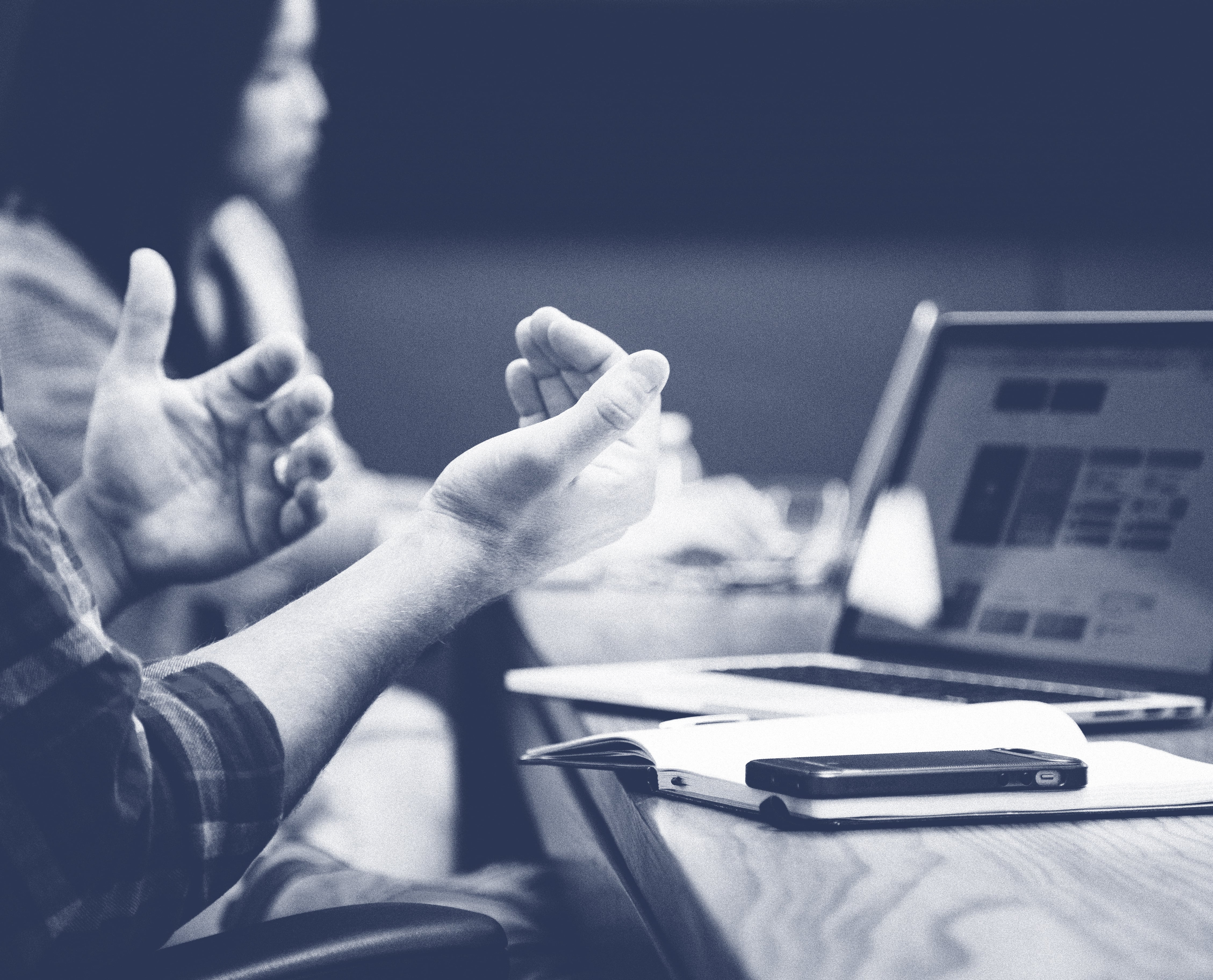 Person gesturing with hands while sitting at a desk with an open laptop, notebook, and smartphone.