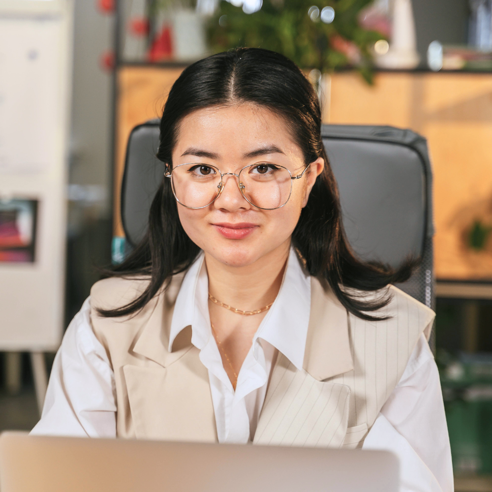 Young woman with glasses sitting at a laptop in an office setting, smiling at the camera.