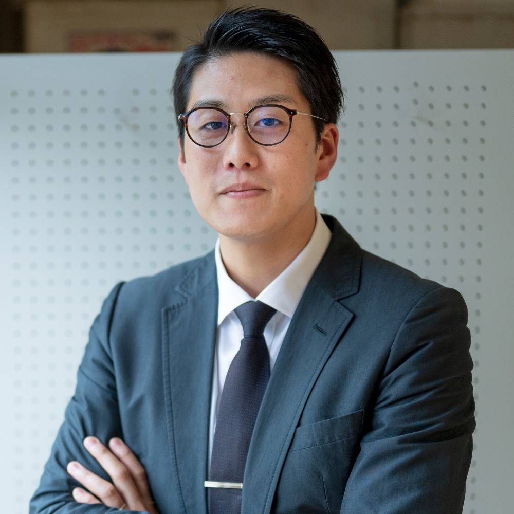 Confident man in glasses and a dark suit with arms crossed standing in front of a perforated panel.