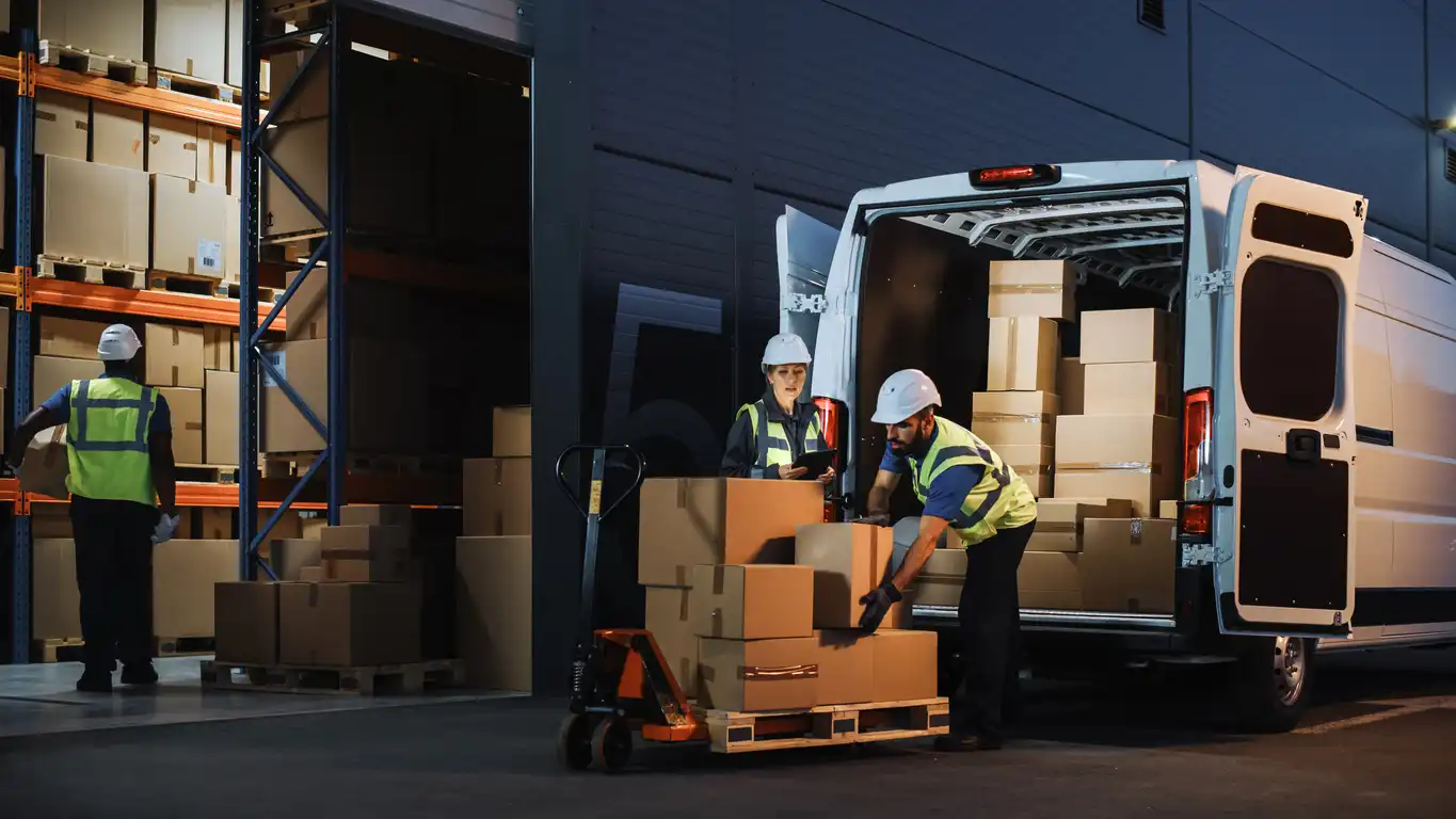 Warehouse workers loading cardboard boxes into a white delivery van at night.