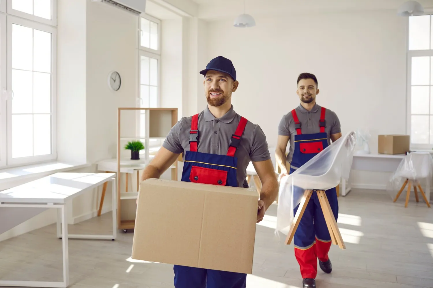 Two movers in matching uniforms carrying a box and a wrapped chair in a bright modern office.