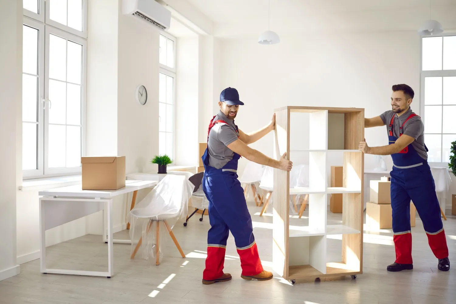 Two professional movers in blue and red uniforms carefully relocating a wooden bookshelf in a bright, modern workplace, providing expert office mover services.