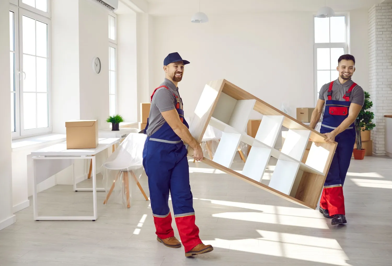 Two professional movers in blue and red uniforms work together to lift and carry a wooden shelving unit in a sunlit, modern workspace, providing expert office mover services.