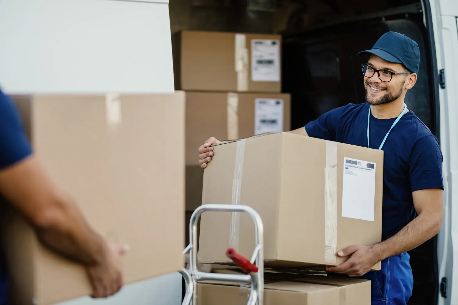 Smiling mover in blue uniform holding a cardboard box near a delivery van with stacked packages.