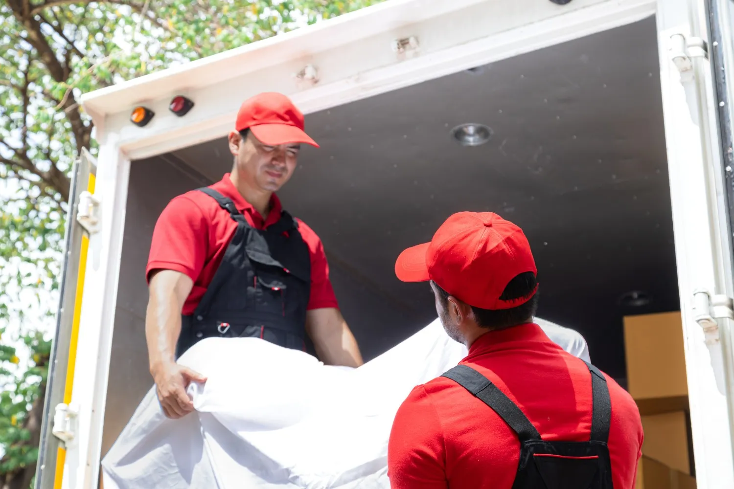 Professional piano movers in red uniforms carefully loading a large, white-wrapped item into the back of a moving truck.