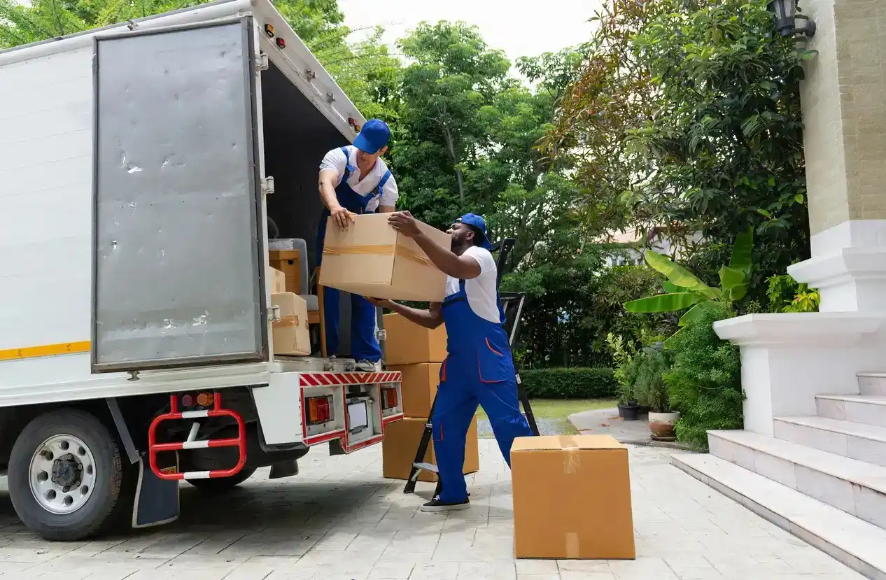 Two movers in blue overalls unloading cardboard boxes from the back of a white truck in a residential driveway.
