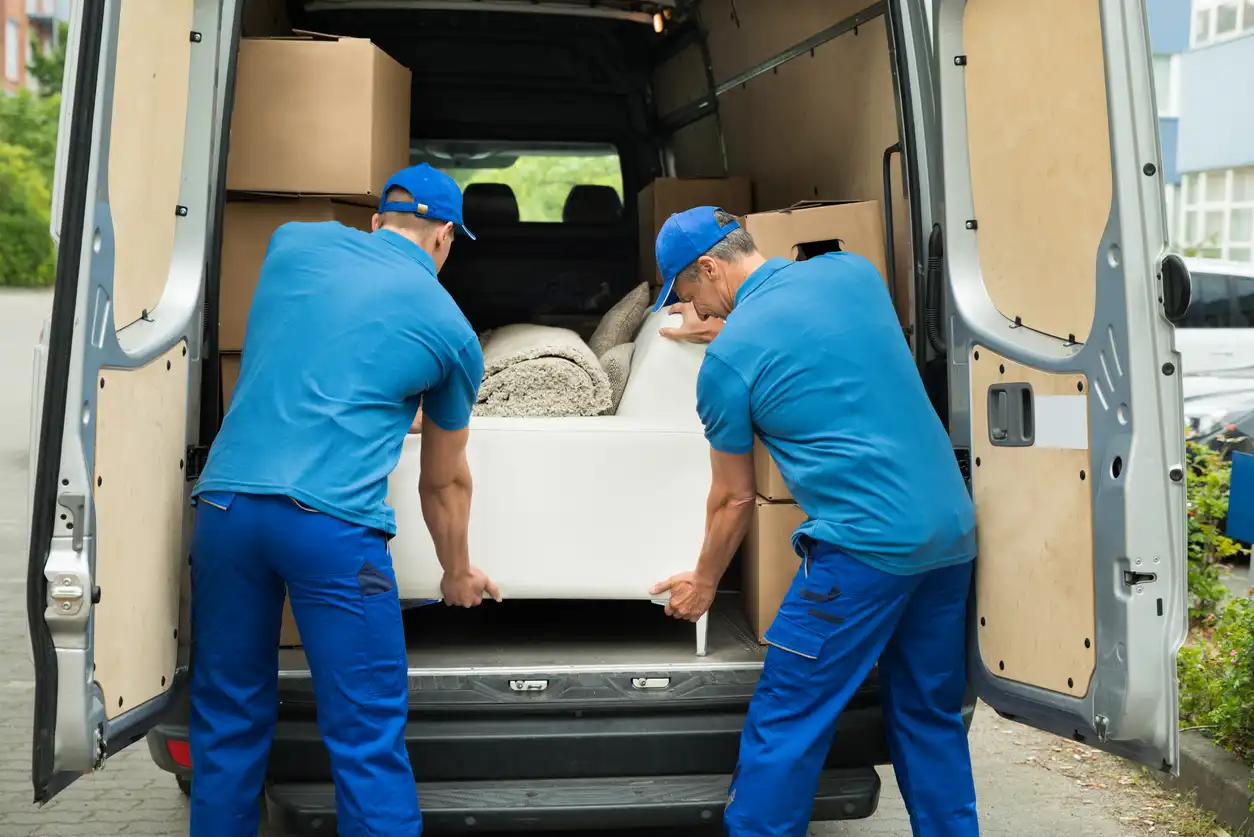 Two movers in blue uniforms loading a white sofa and rolled-up rugs into the back of a van.