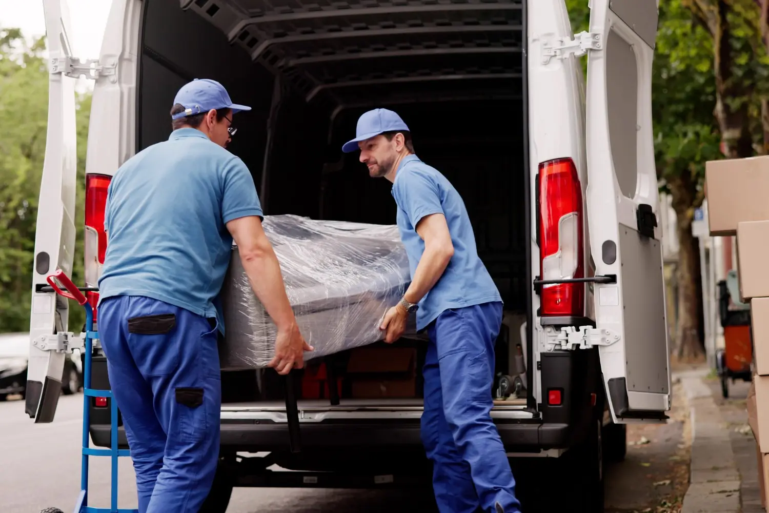 Two movers in blue uniforms loading a wrapped furniture piece into a white cargo van with boxes nearby.
