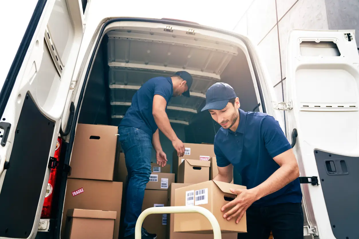 Two delivery men in blue shirts and caps organize cardboard boxes inside a white van