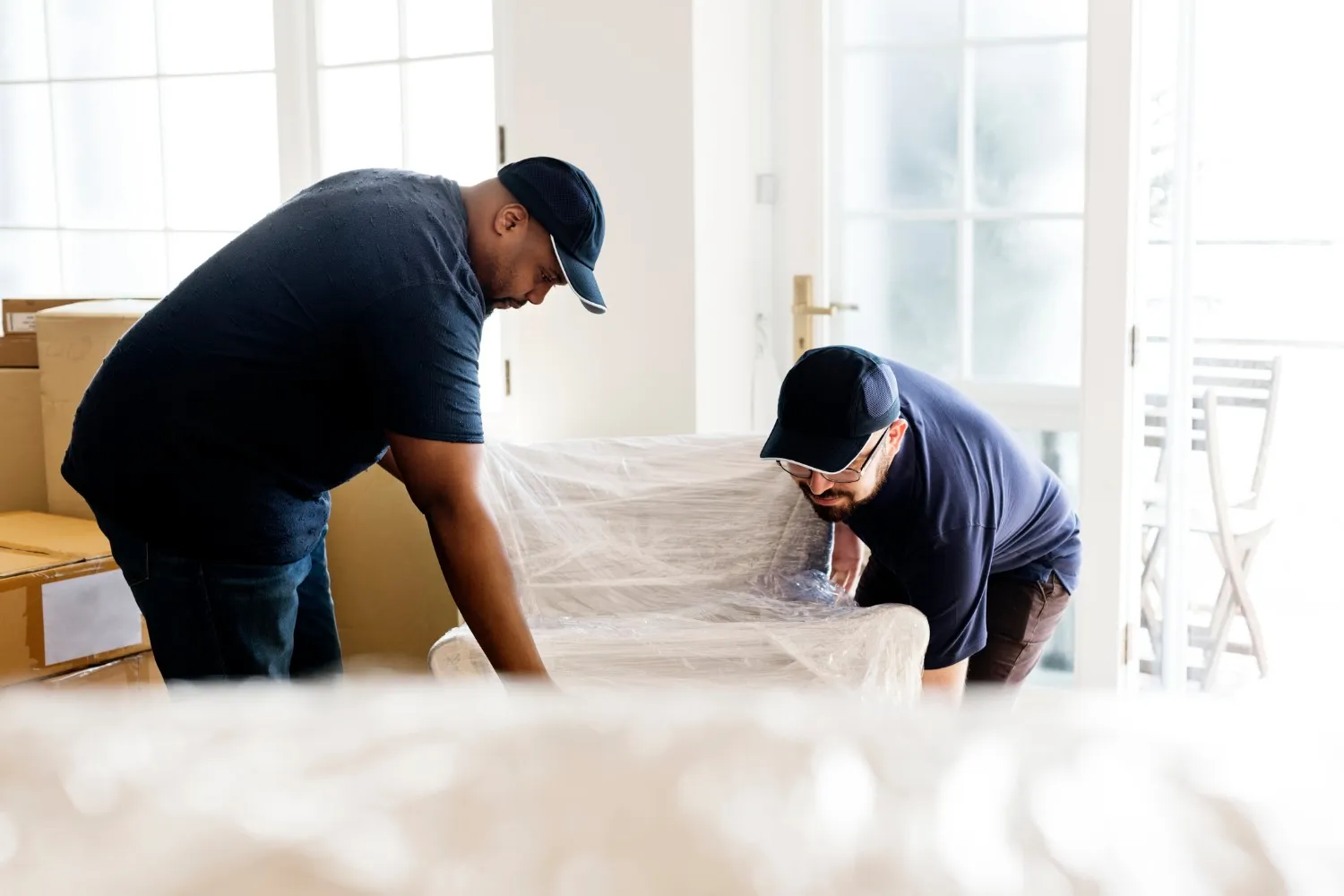 Two individuals in dark shirts and caps lifting a plastic-wrapped cushioned chair indoors, with boxes nearby.