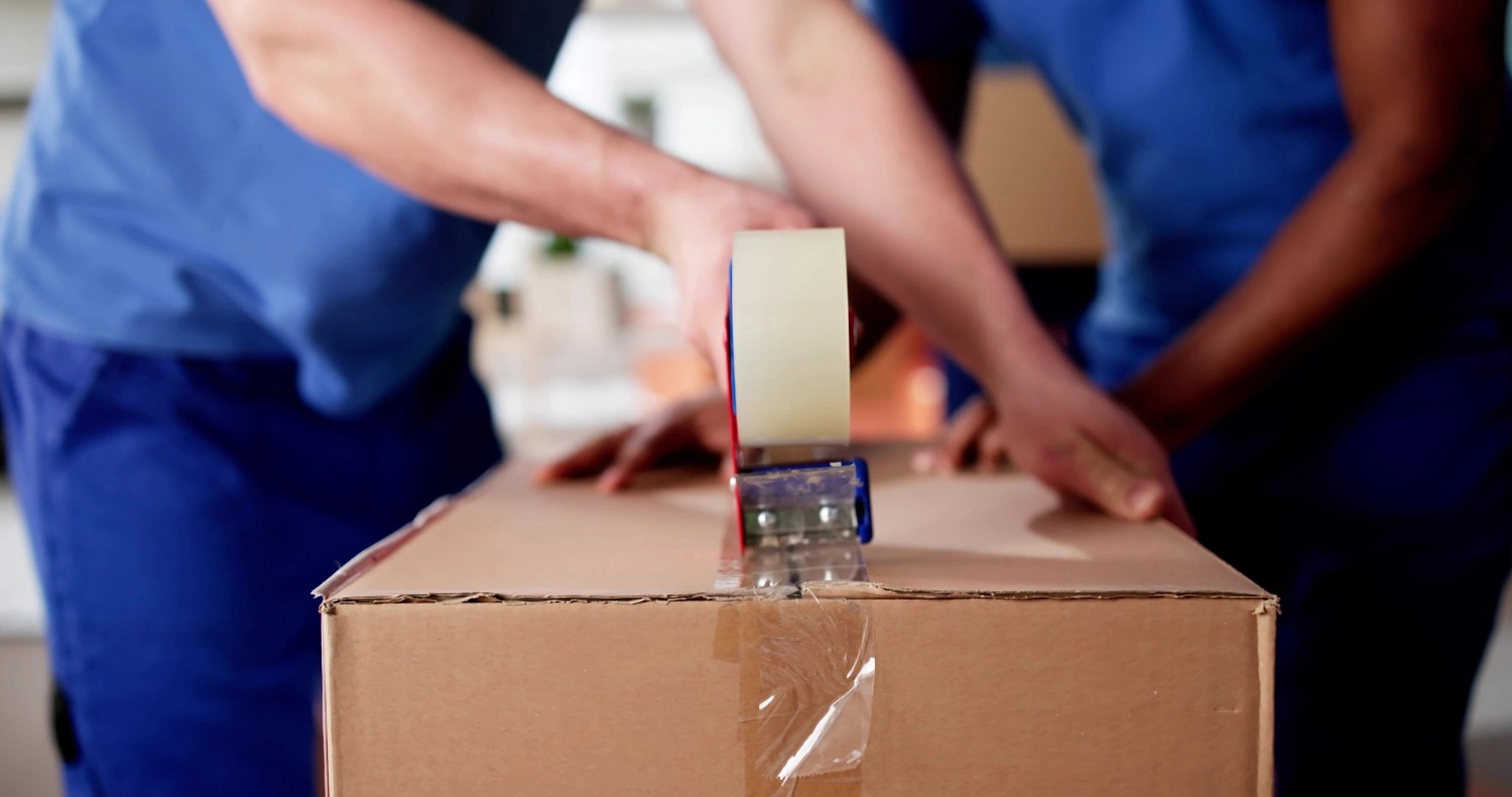 Two individuals in blue uniforms packing cardboard boxes indoors, with shelves and a third person in background.