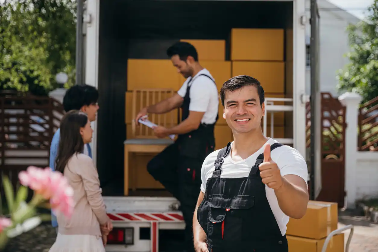 A smiling mover giving a thumbs-up in front of a truck filled with boxes while a couple stands nearby.