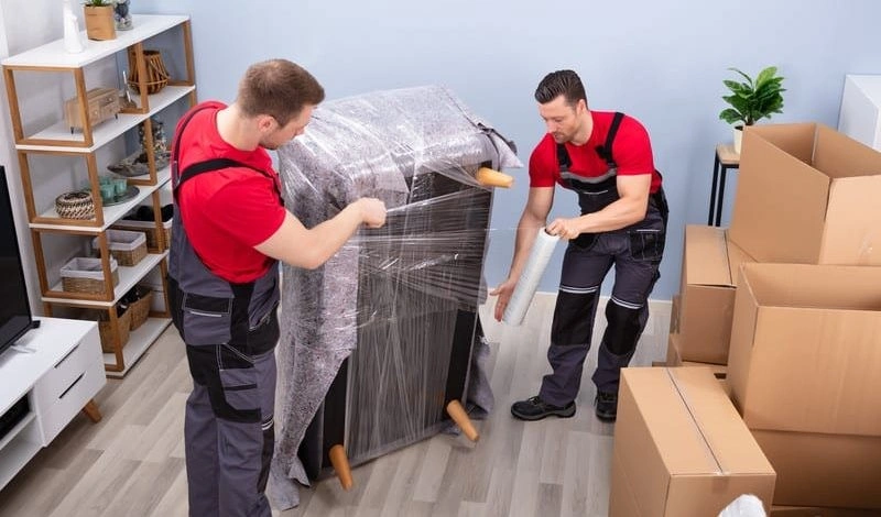 Two movers wrapping furniture in plastic.