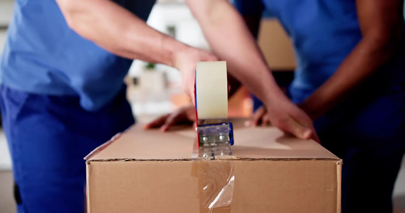 A detailed close-up of a worker's hands using a tape dispenser to secure a box