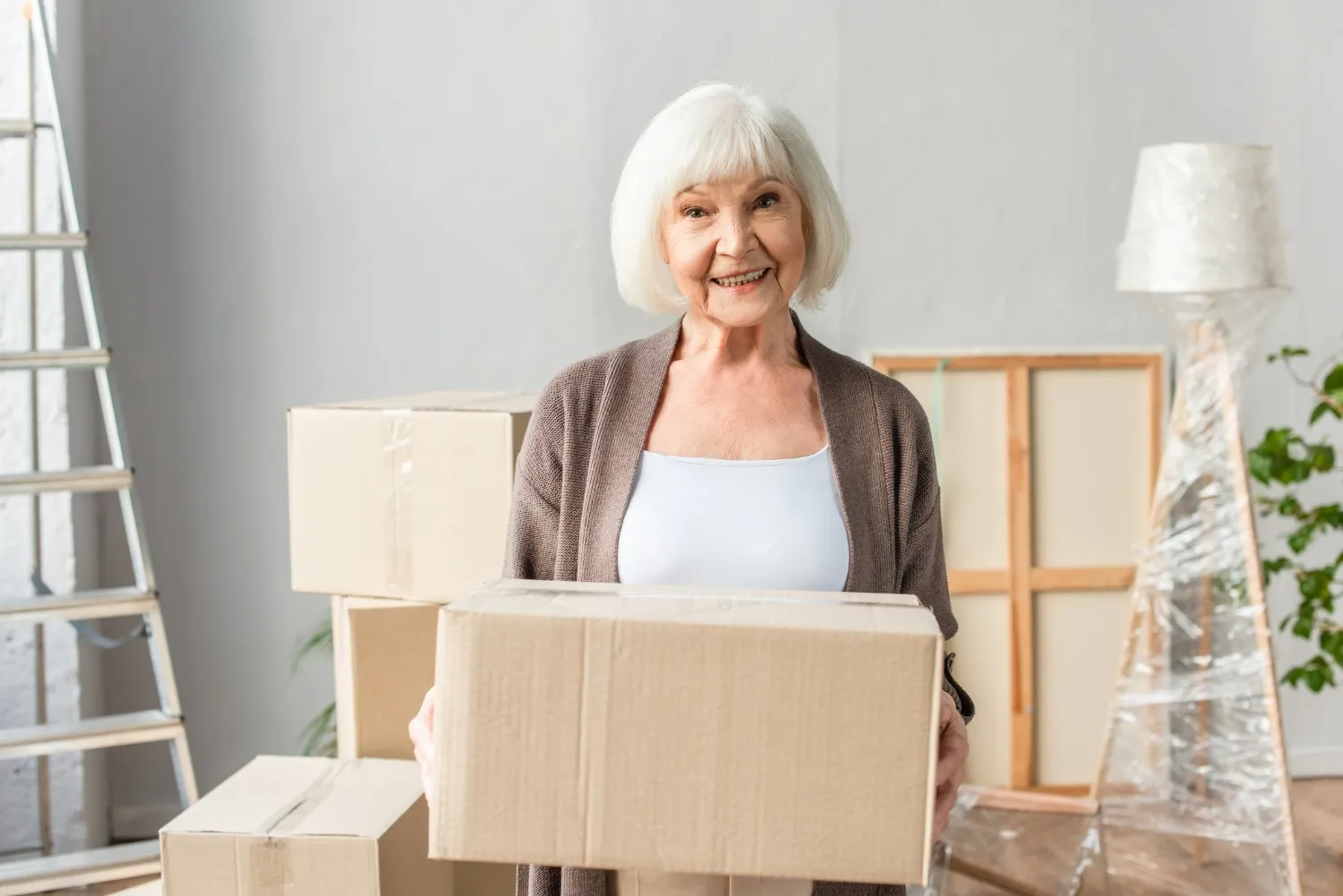 A smiling elderly woman holding a cardboard box while utilizing senior moving services in a room filled with packed items and moving equipment.