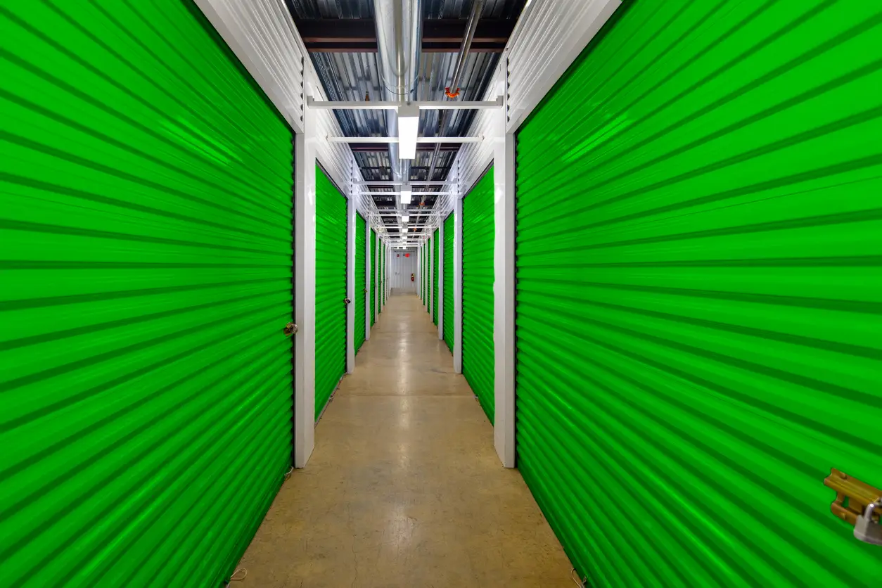 A long, brightly lit indoor corridor featuring rows of vibrant green corrugated metal storage unit doors