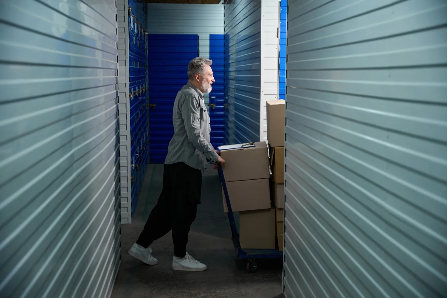 Person in gray shirt pushing a cart with cardboard boxes down a corridor of blue storage unit doors.