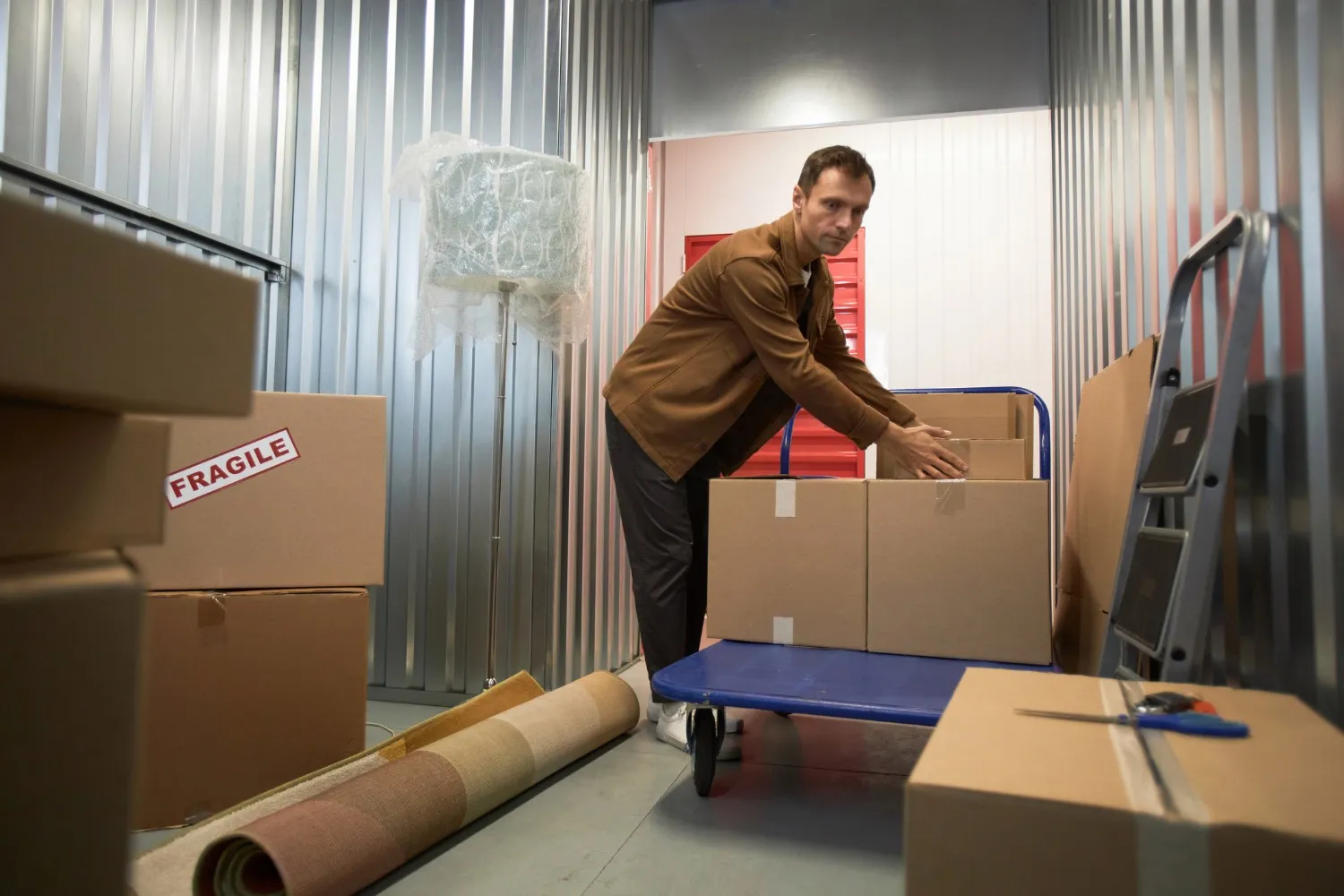 Person organizing cardboard boxes on a blue dolly inside a storage unit with a rug and lamp nearby.