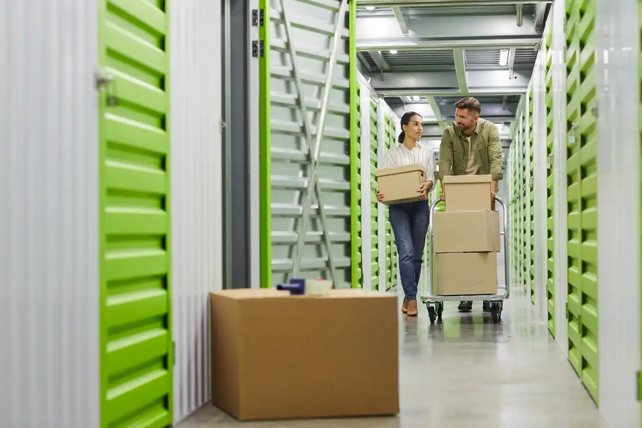 A man and woman moving boxes on a trolley through a storage facility hallway.