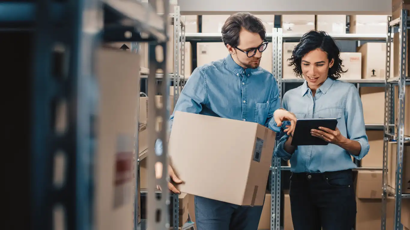 Two colleagues reviewing information on a tablet while holding a box in a warehouse.