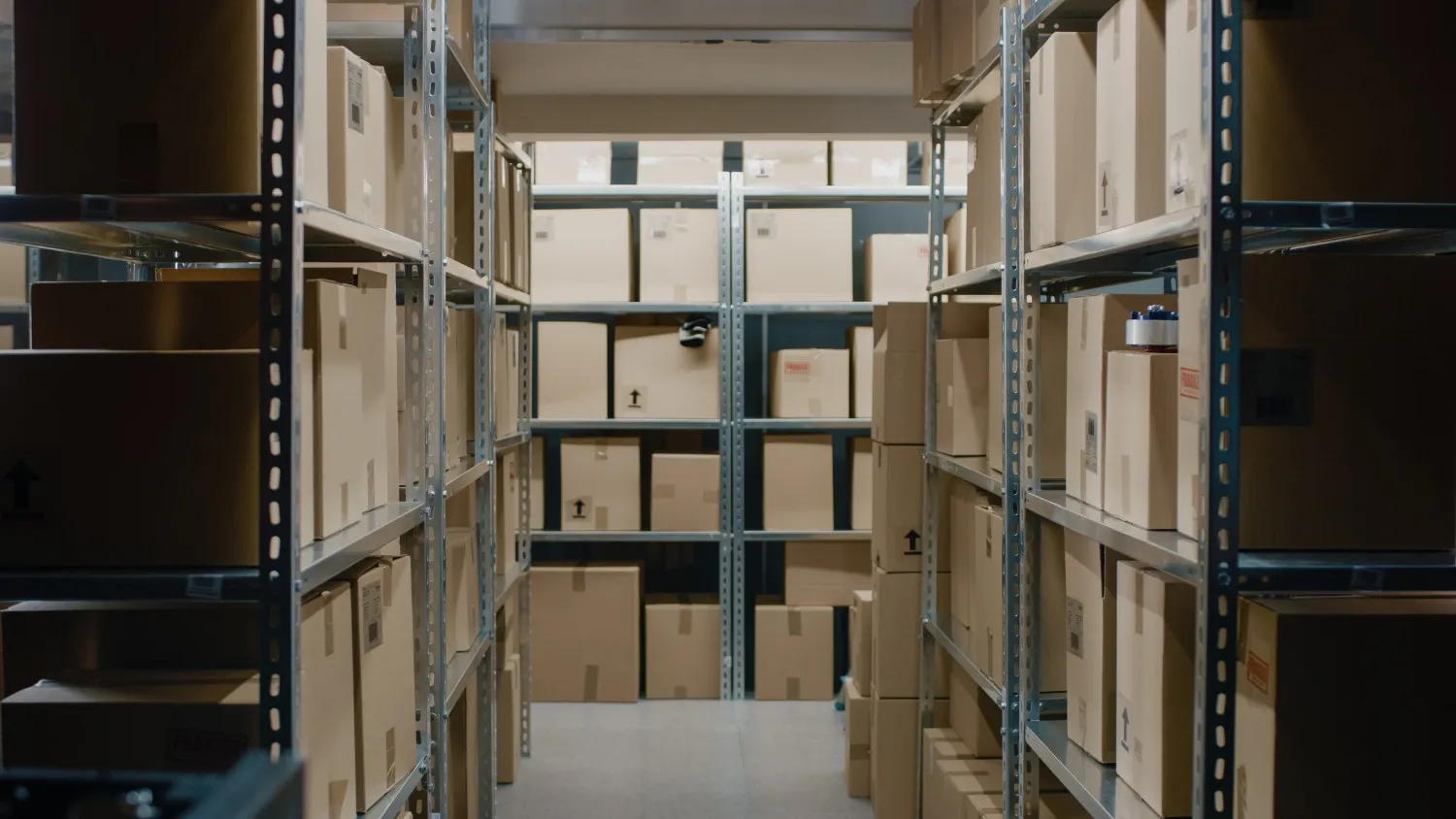 Metal shelving units filled with labeled cardboard boxes in a brightly lit warehouse or storage room.