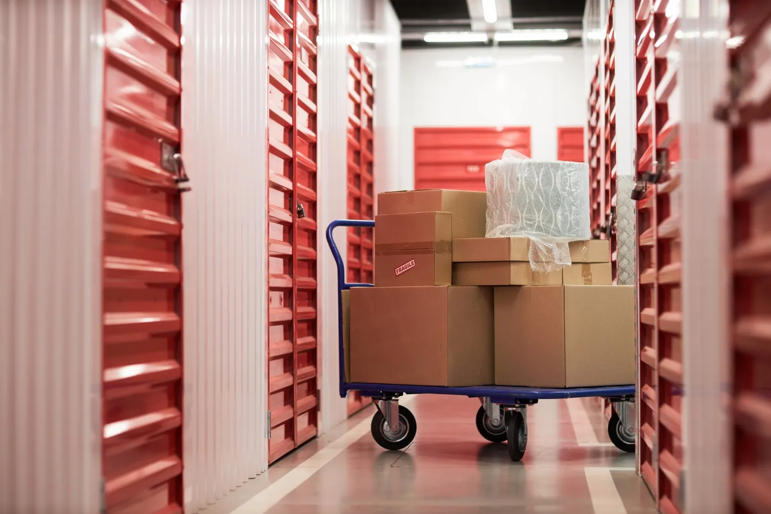 Blue cart loaded with cardboard boxes and bubble wrap in a hallway of red storage unit doors.