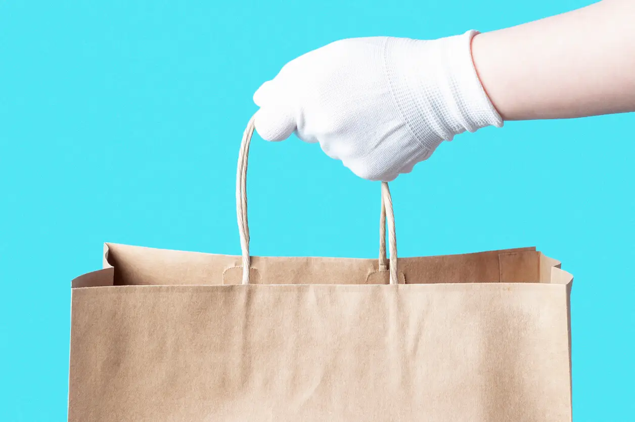 A close-up shot of a hand in a white glove holding a brown paper shopping bag