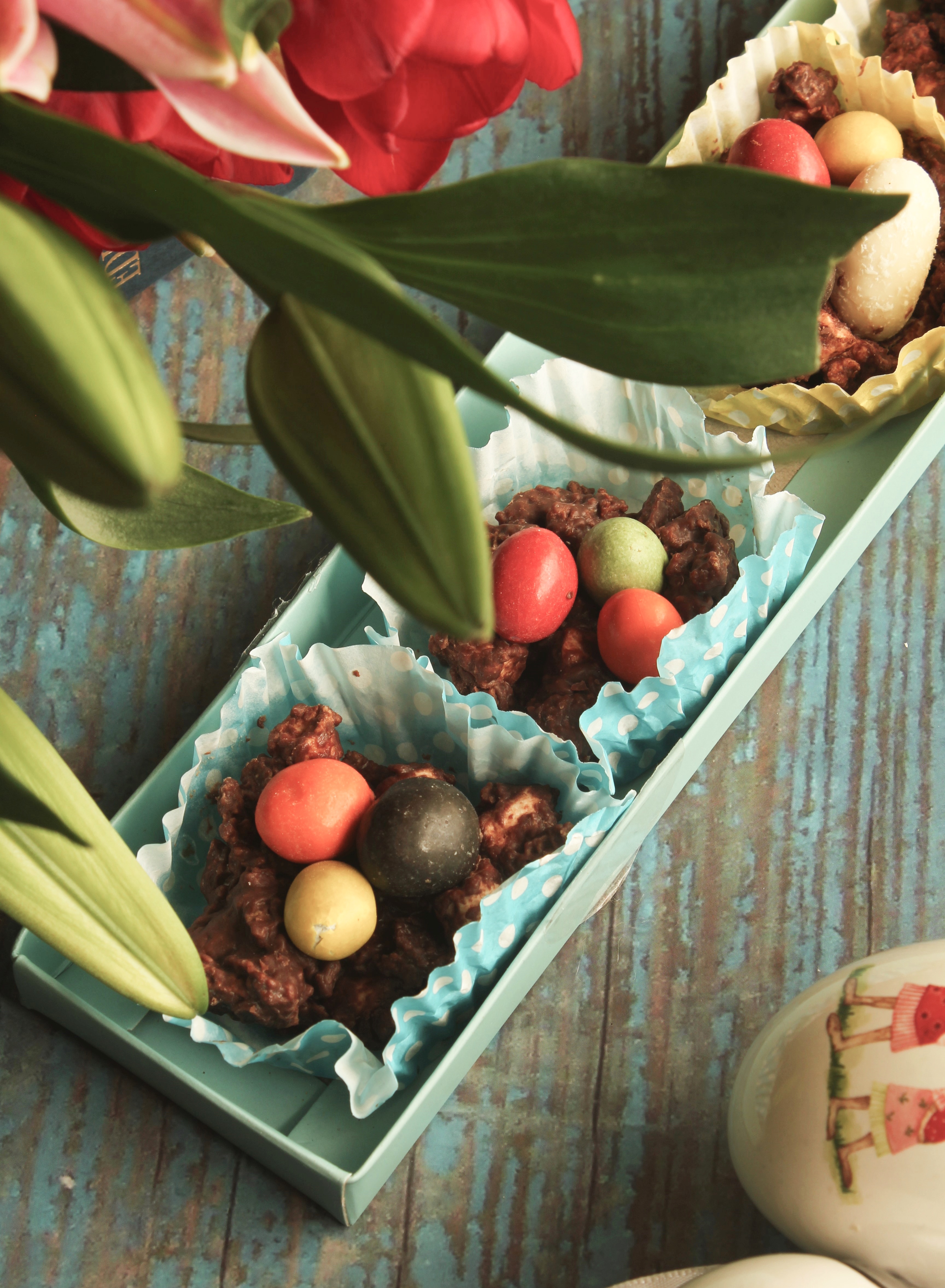 Close-up of colorful chocolate candies and clusters in blue and yellow polka-dotted paper cups inside a turquoise tray on a rustic blue wooden surface with green leaves and painted eggs nearby.