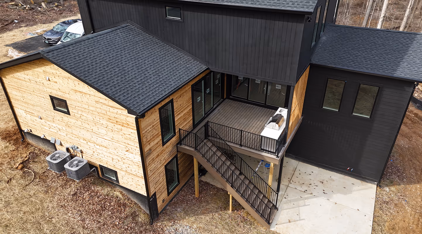 Modern two-story house with black and natural wood siding, elevated deck with stairs, and surrounding yard.