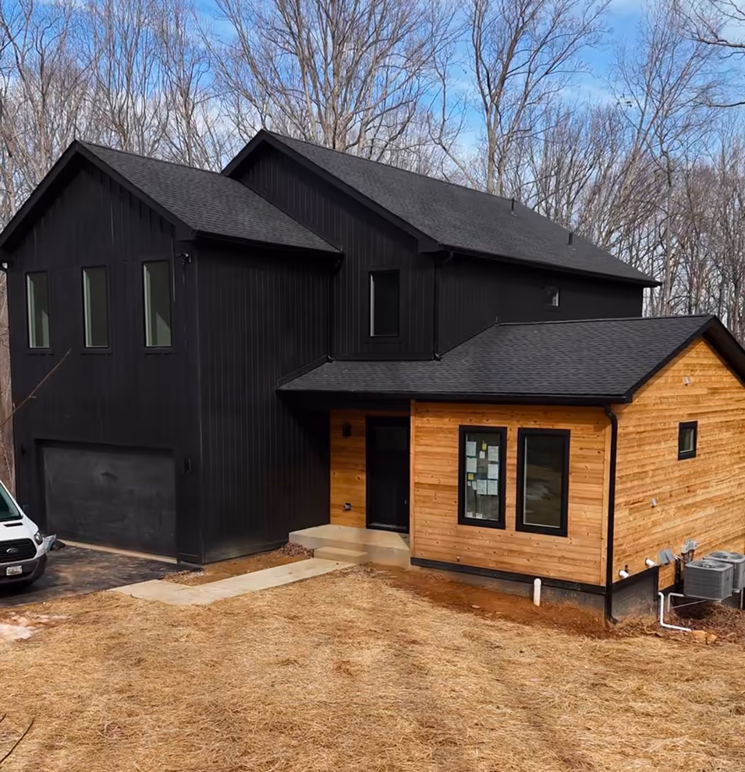 Modern two-story house with black and natural wood exterior siding, set in a wooded area with bare trees.