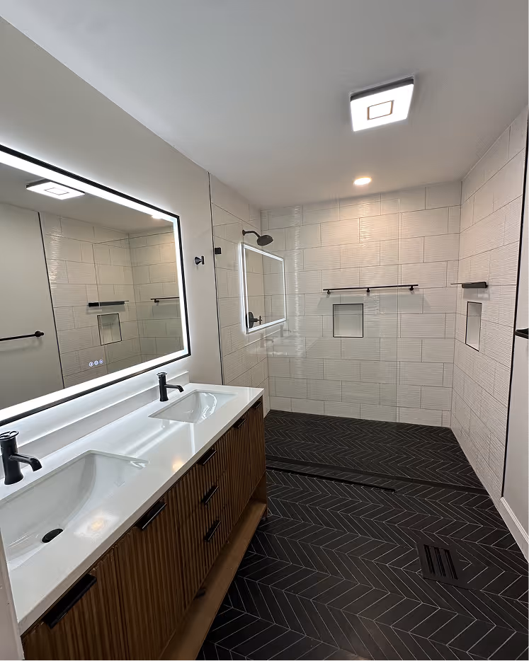 Modern bathroom with dual sinks on a wooden vanity, large illuminated mirror, walk-in shower with black fixtures, white tiled walls, and black herringbone floor tiles.