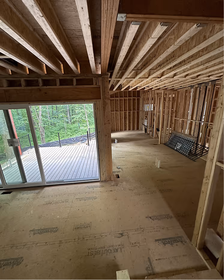Interior view of a wooden house under construction showing exposed ceiling beams, wall framing, and a sliding glass door leading to a wooden deck with greenery outside.
