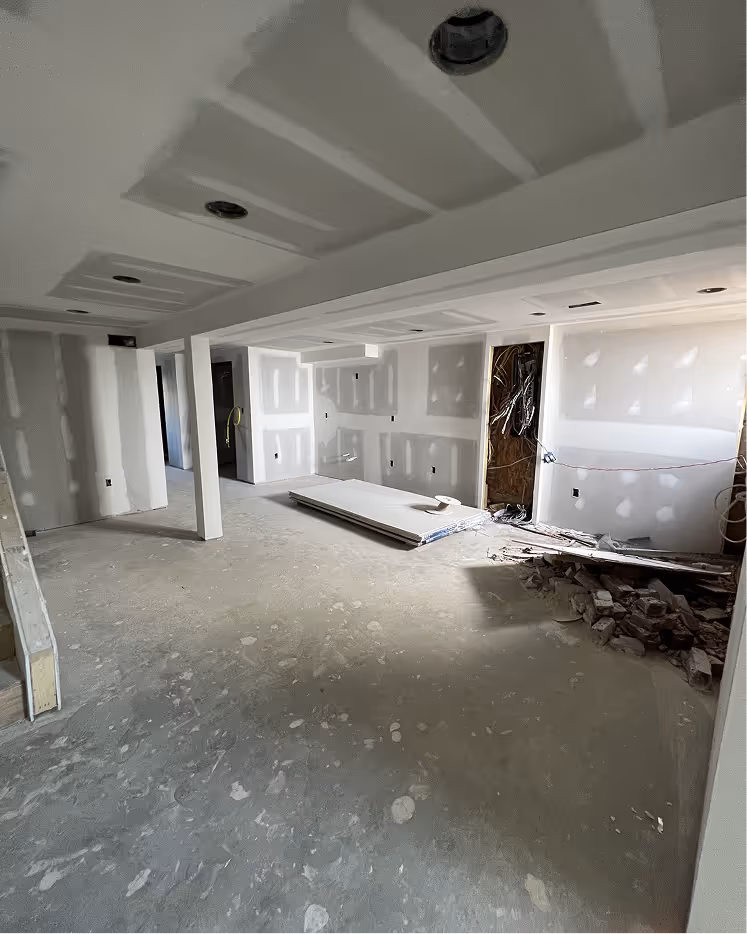 Interior of an unfinished room under construction with drywall installed and cement floor, with building materials and debris on the floor.