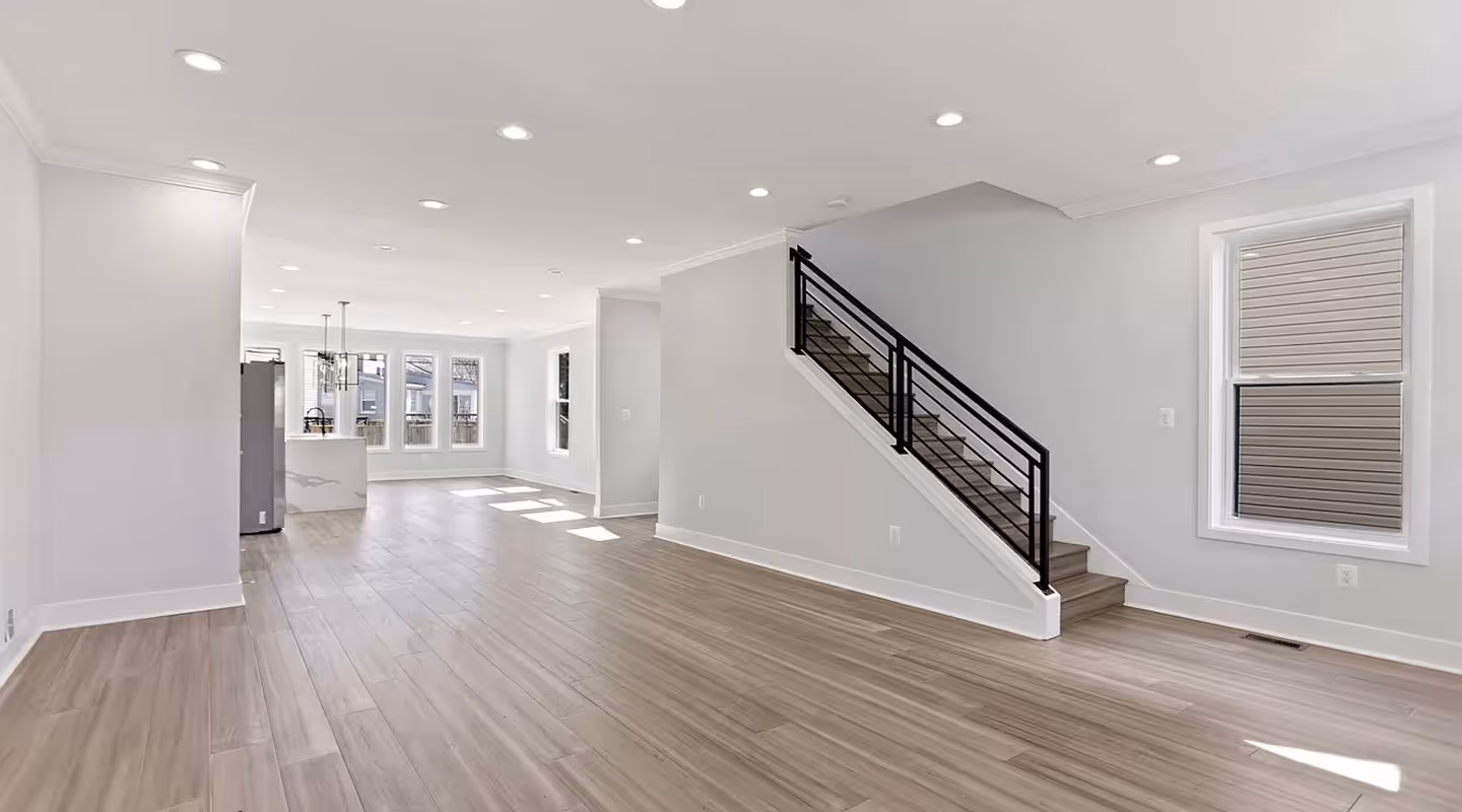 Bright, modern open-concept living space with light wood flooring, white walls, recessed ceiling lights, and a staircase with black metal railing.