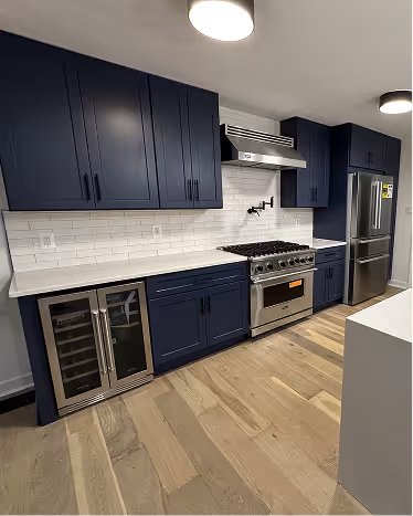 Modern kitchen with navy blue cabinets, stainless steel appliances including a stove, wine cooler, and refrigerator, white subway tile backsplash, and light wood flooring.