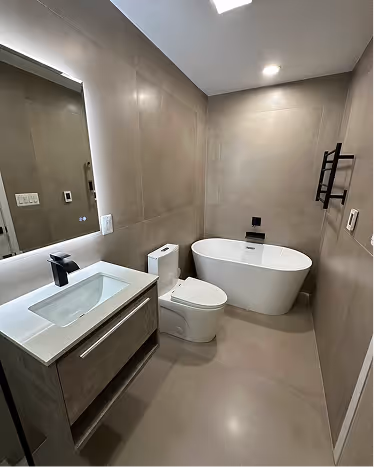 Modern bathroom with grey walls featuring a white bathtub, a toilet, and a vanity with a sink and illuminated mirror.
