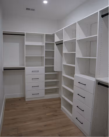 Empty walk-in closet with white built-in shelves, drawers, hanging rods, and wood flooring.