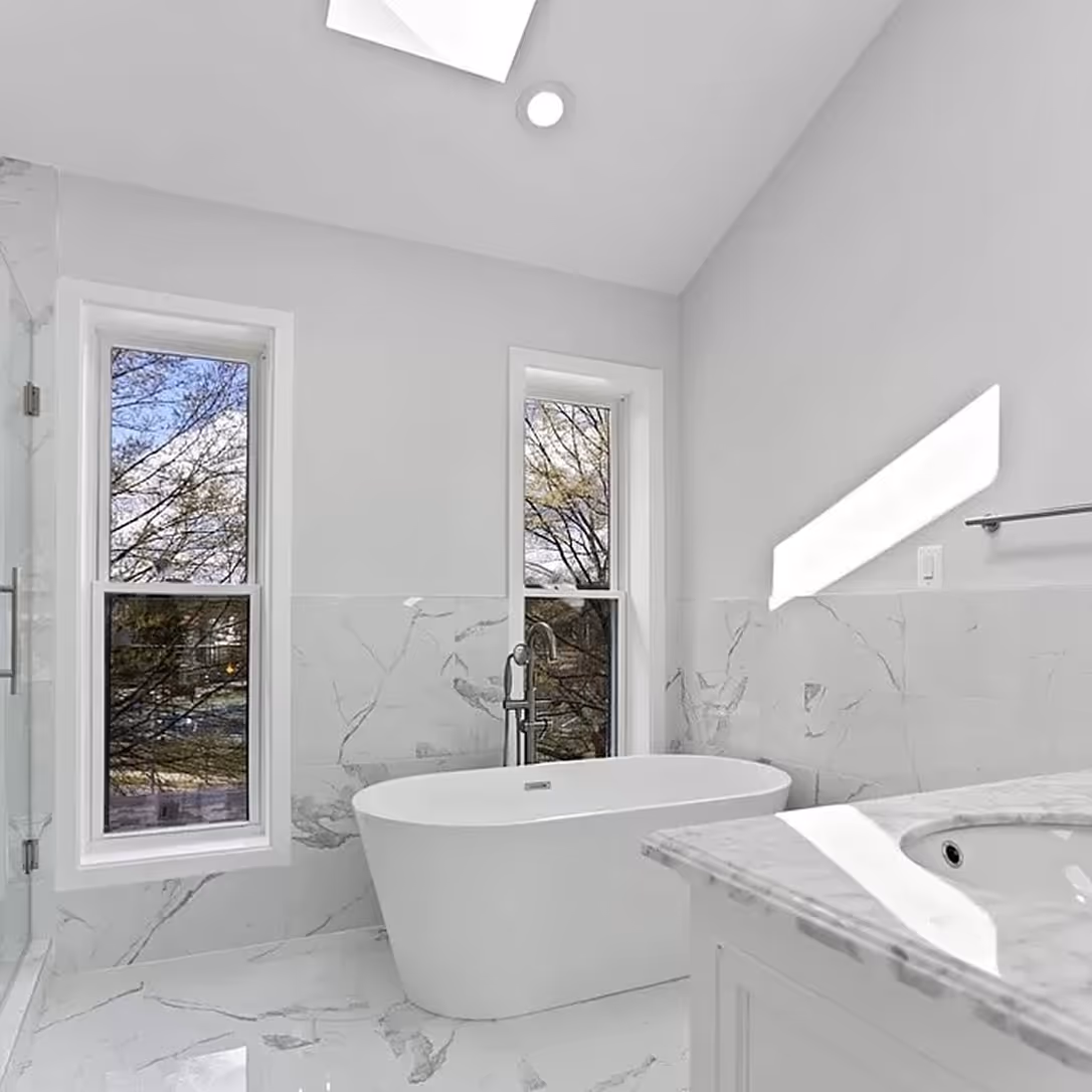 Modern bathroom with white freestanding tub, marble tiled floor and walls, two tall windows, and a marble countertop sink.