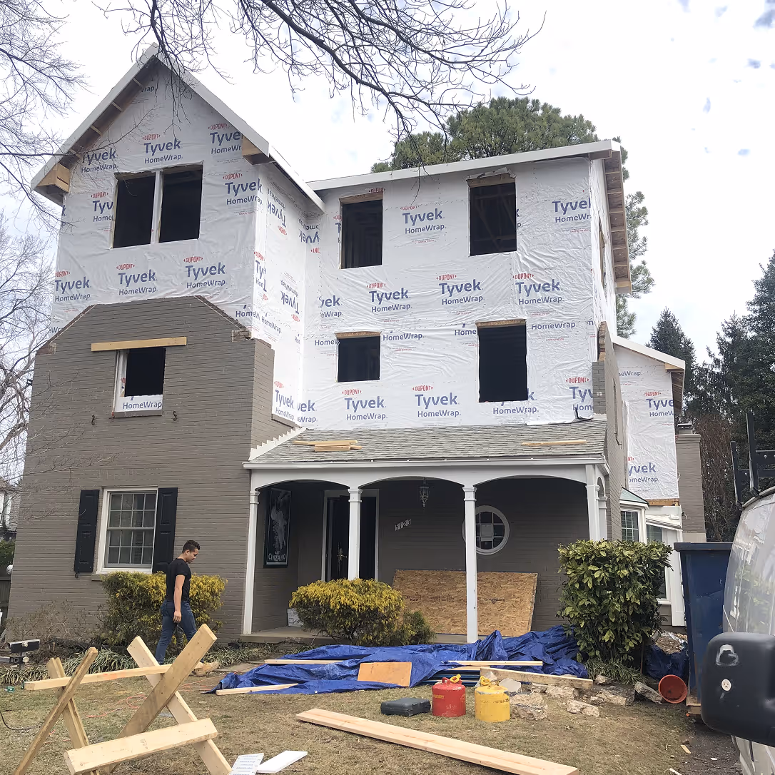 Two-story house under renovation with Tyvek HomeWrap on upper floors, construction materials on grass, and a person walking near the entrance.