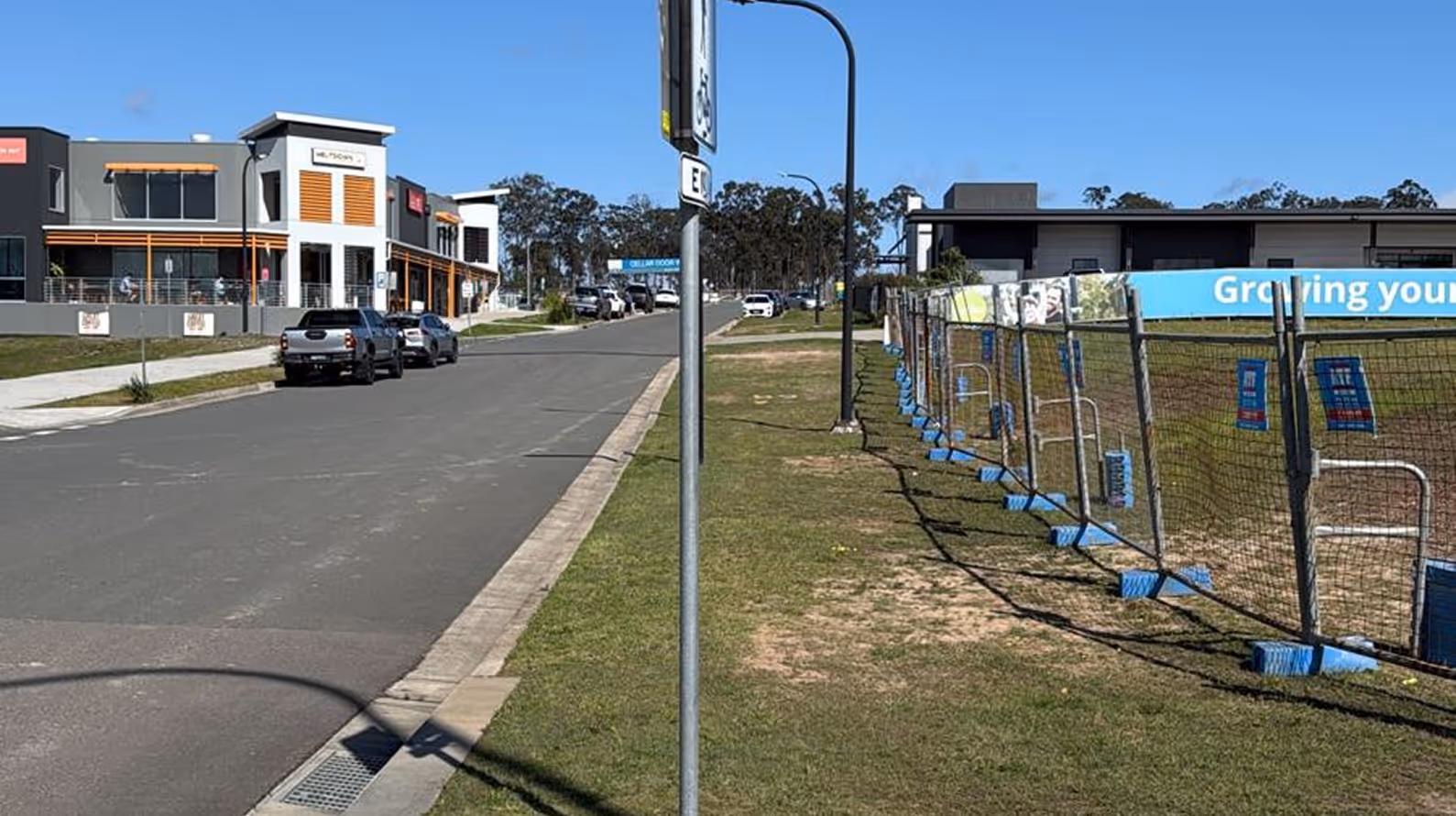 Suburban street with parked cars beside modern commercial buildings on left and temporary fencing along a grassy area on right under a clear blue sky.