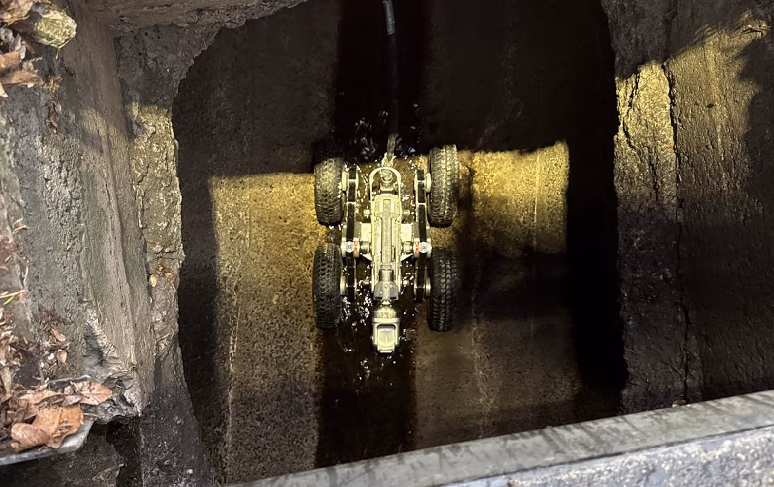 Top-down view of a small robotic vehicle with six wheels inside a narrow, dark concrete shaft or tunnel.