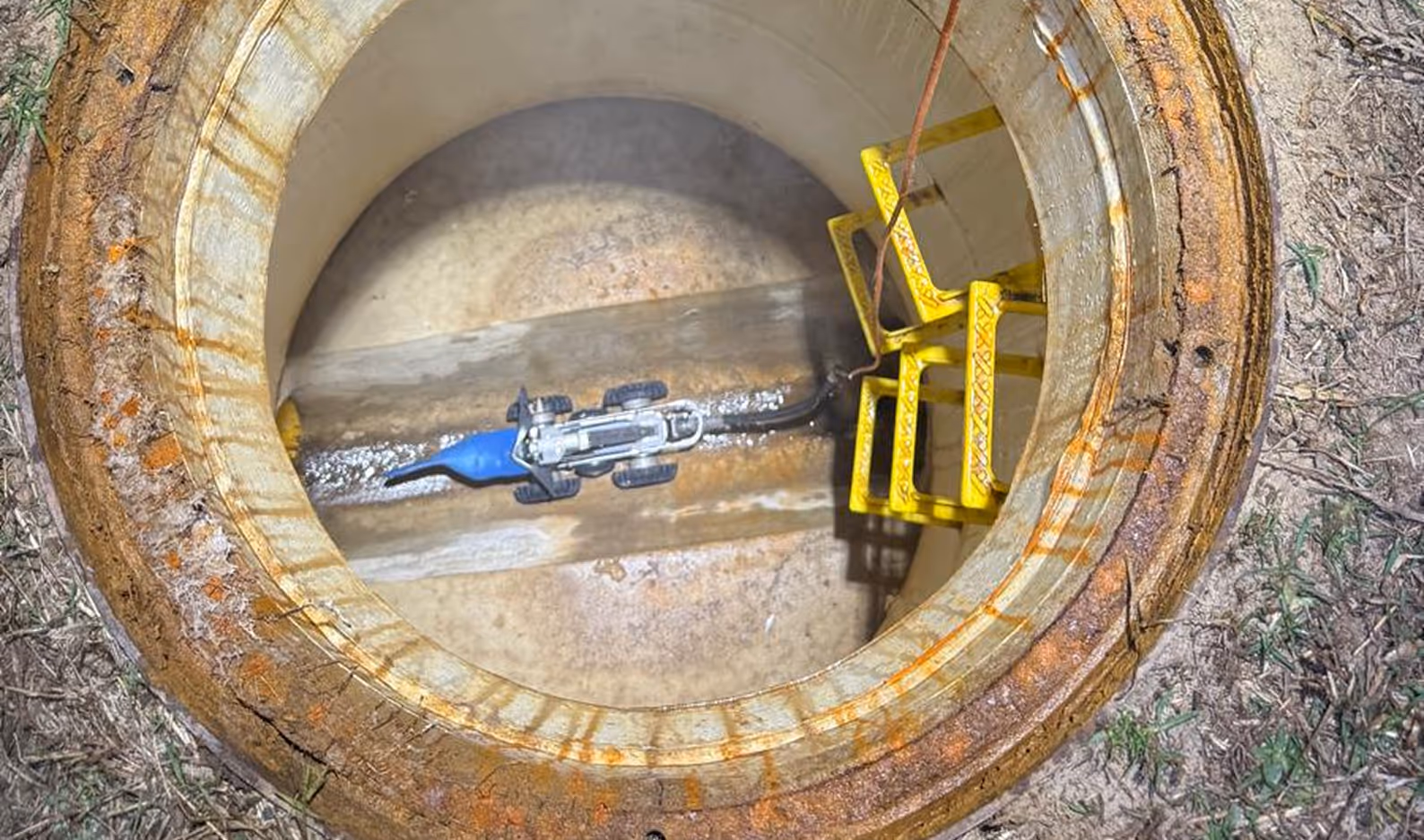 View down into a cylindrical concrete manhole with a yellow metal ladder and a blue robotic inspection device inside.