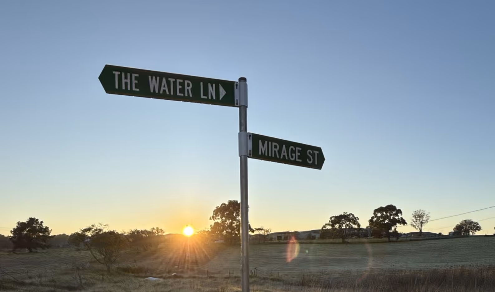 Street signs for The Water Ln and Mirage St on a pole against a sunrise over a rural landscape.