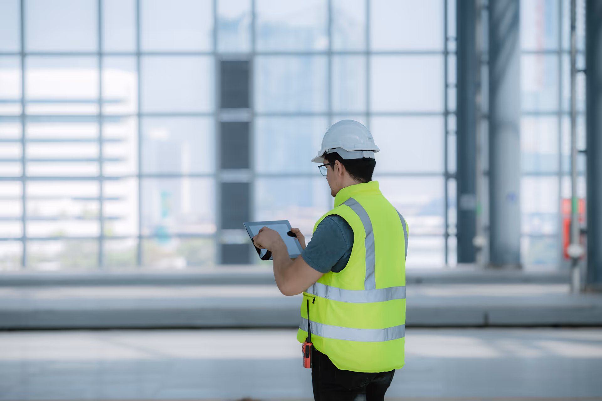 Construction worker wearing a white hard hat and neon yellow safety vest using a tablet inside a building under construction.
